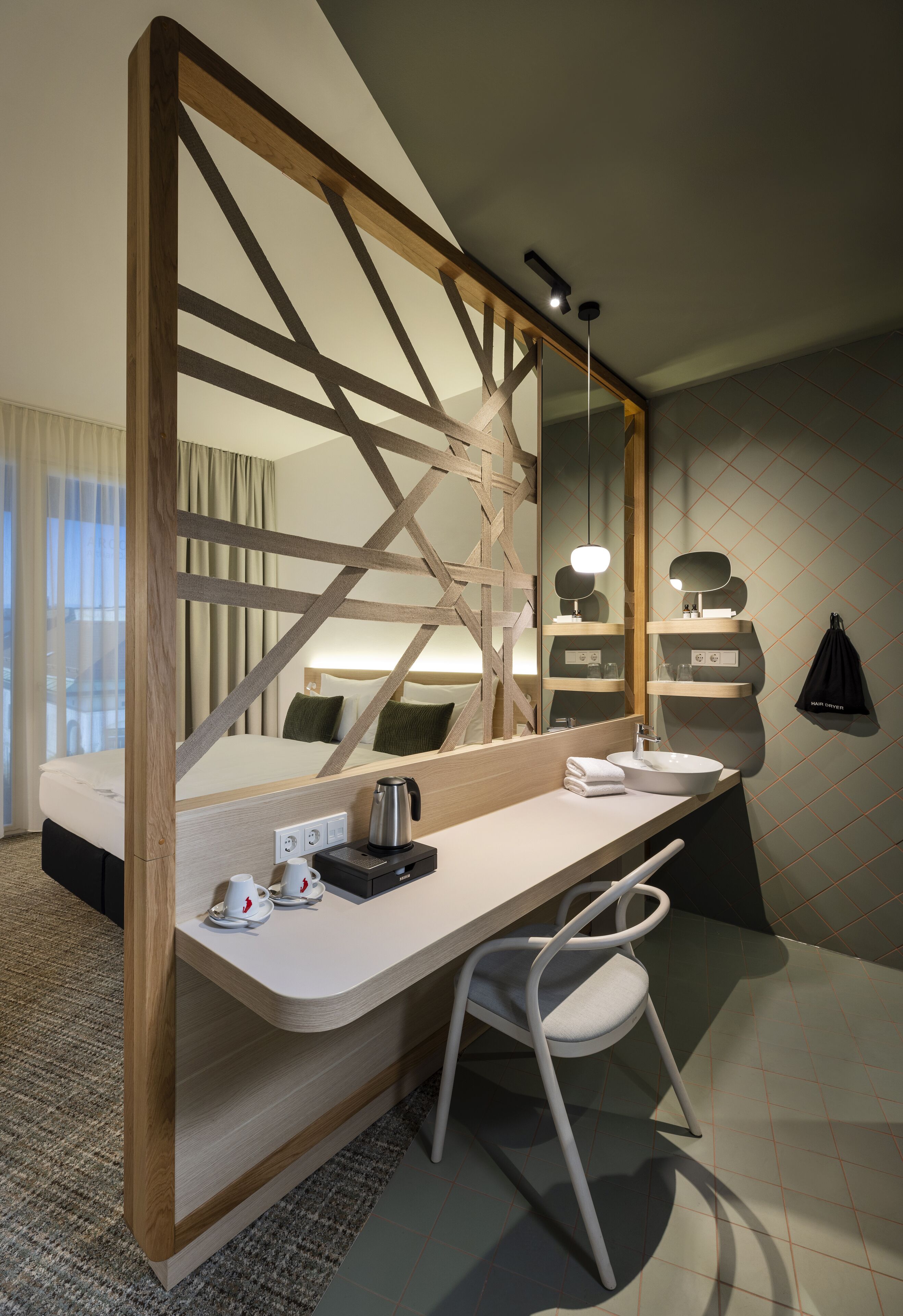 A modern hotel room with a wooden-framed bed, a large mirror, a white desk with a kettle, cups, and a black bag hanging on the wall.