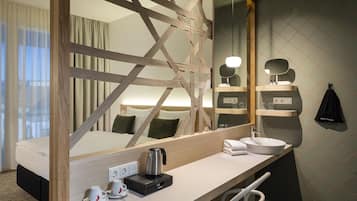 A modern hotel room with a wooden-framed bed, a large mirror, a white desk with a kettle, cups, and a black bag hanging on the wall.