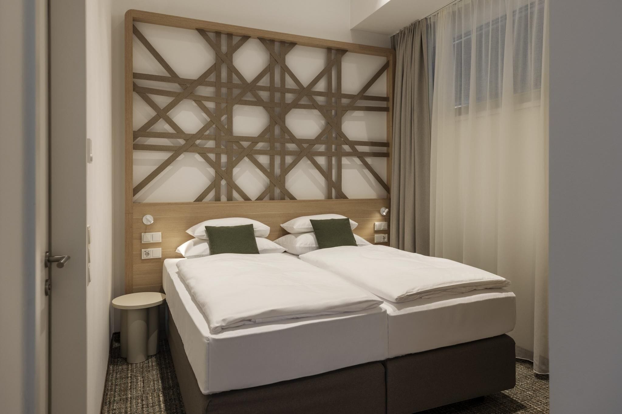A hotel room with a double bed, a wooden headboard with a geometric pattern, a bedside table, and a window with sheer curtains.