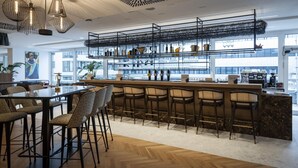 A modern bar area with a wooden herringbone floor, black metal shelving with glass doors, and a variety of bar stools. - ARCOTEL AQ Wien (Vienna)