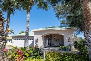 A house with a white garage door, a covered porch, and a well-maintained garden with various plants and a palm tree.