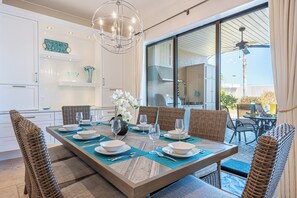 A dining area with a large table set for six, surrounded by wicker chairs, a chandelier, and a view of the outdoors.
