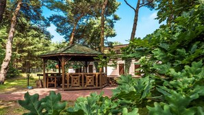Una pérgola con techo y estructura de madera, rodeada de árboles y un sendero de ladrillos.