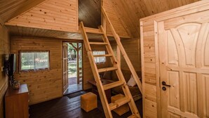 Interior de una cabaña de madera con un entrepiso para dormir, una escalera al entrepiso, una puerta y una ventana.