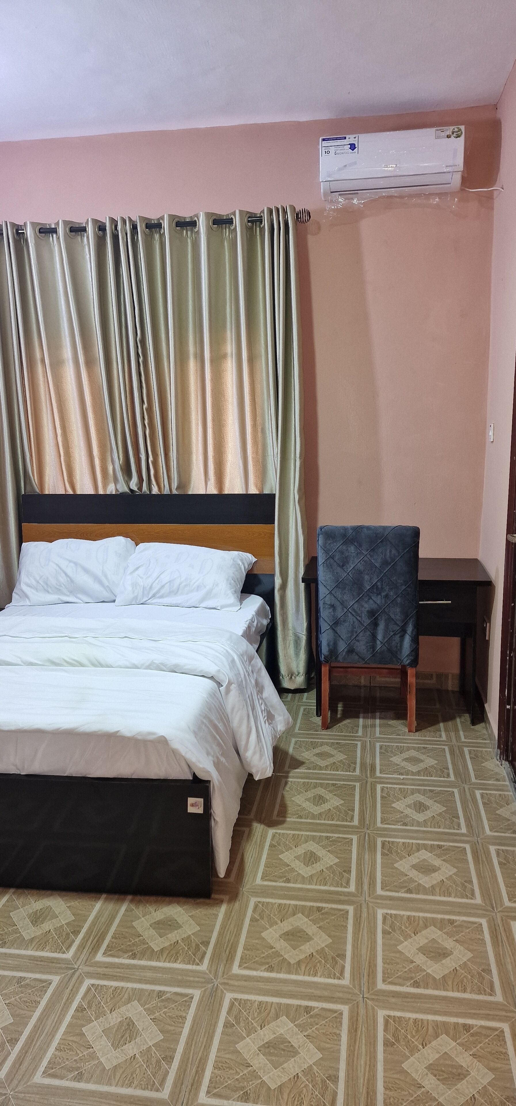 A hotel room with a bed, a desk, a chair, an air conditioner, and patterned flooring.