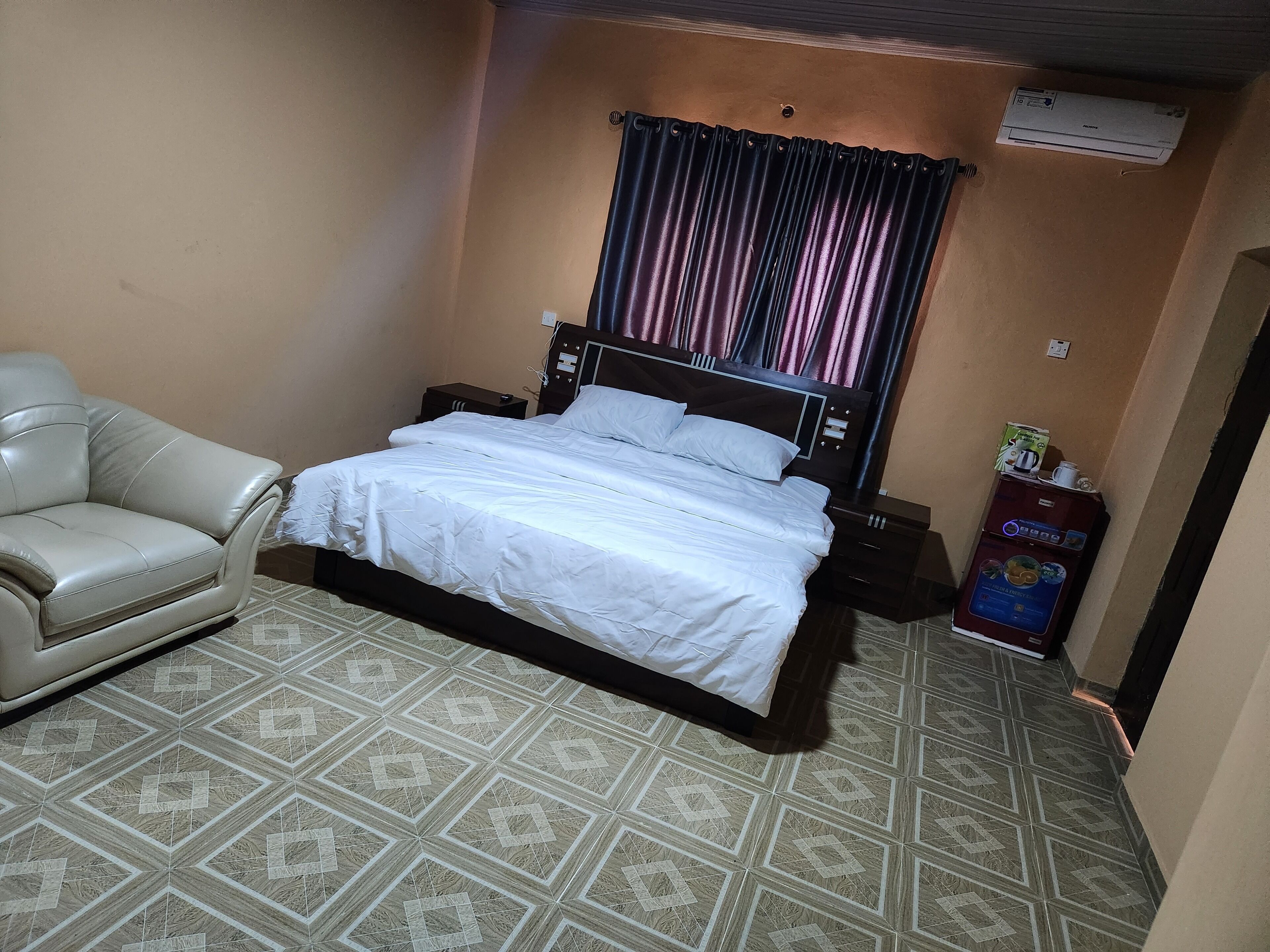 A bedroom with a bed, a chair, a nightstand, an air conditioner, and a coffee machine.