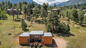 A modern two-story building with a flat roof, surrounded by a forested area with mountains in the background.