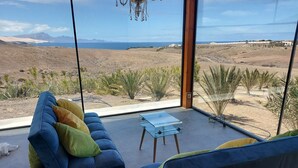 A modern living room with a sofa and a glass coffee table, large windows offering a view of a desert landscape and mountains. - Casa Julie, Ferien in Absoluter Ruhe (La Pared)