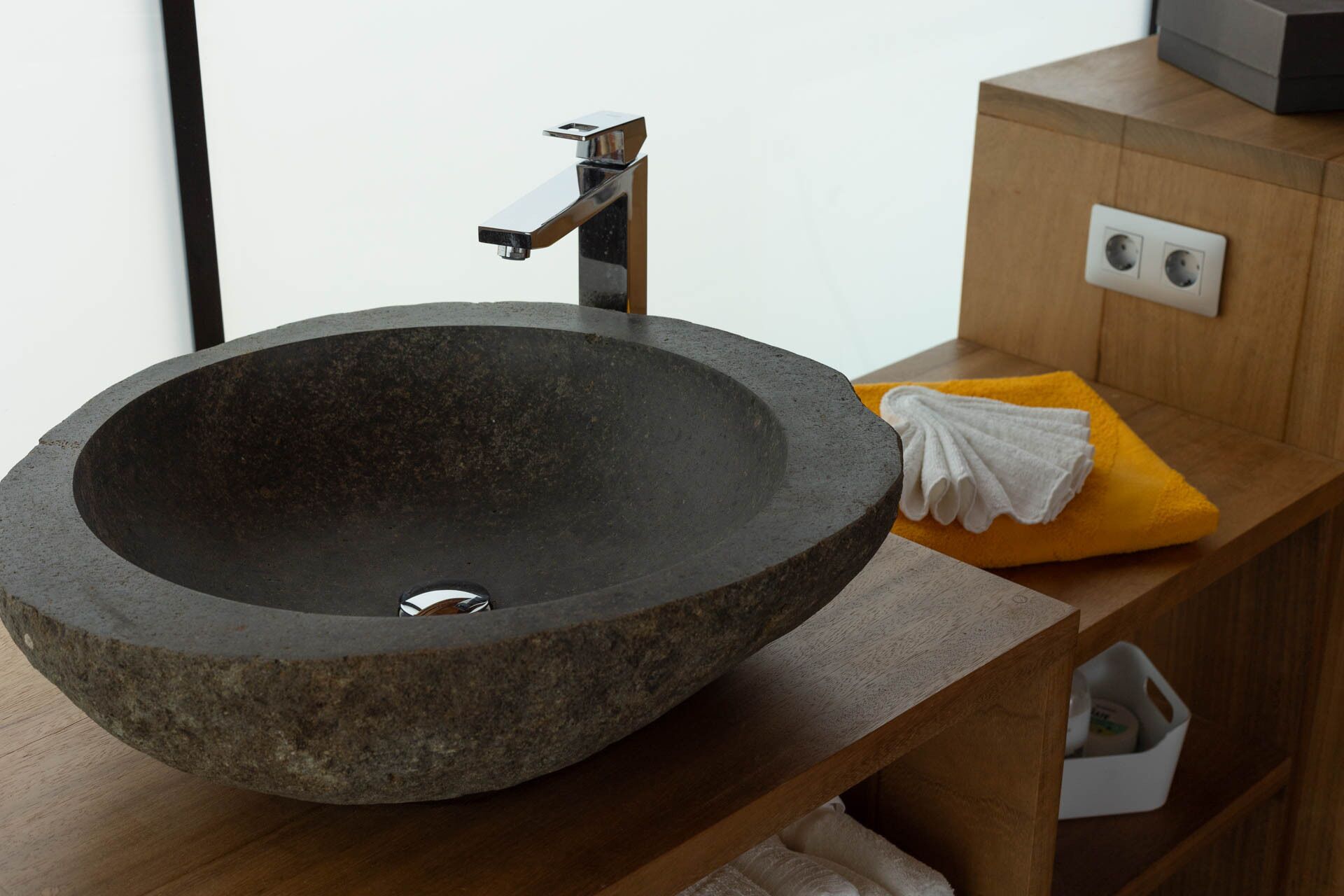 A modern bathroom with a large, dark stone sink, a sleek faucet, and a wooden vanity with a yellow towel and a white towel.