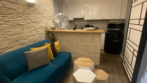 Un ambiente compatto con un divano blu, tavolini in legno, angolo cottura con pensili bianchi e una parete in pietra.