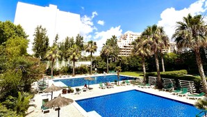 Une grande piscine extérieure entourée de palmiers et de chaises longues, sous un ciel bleu azur.