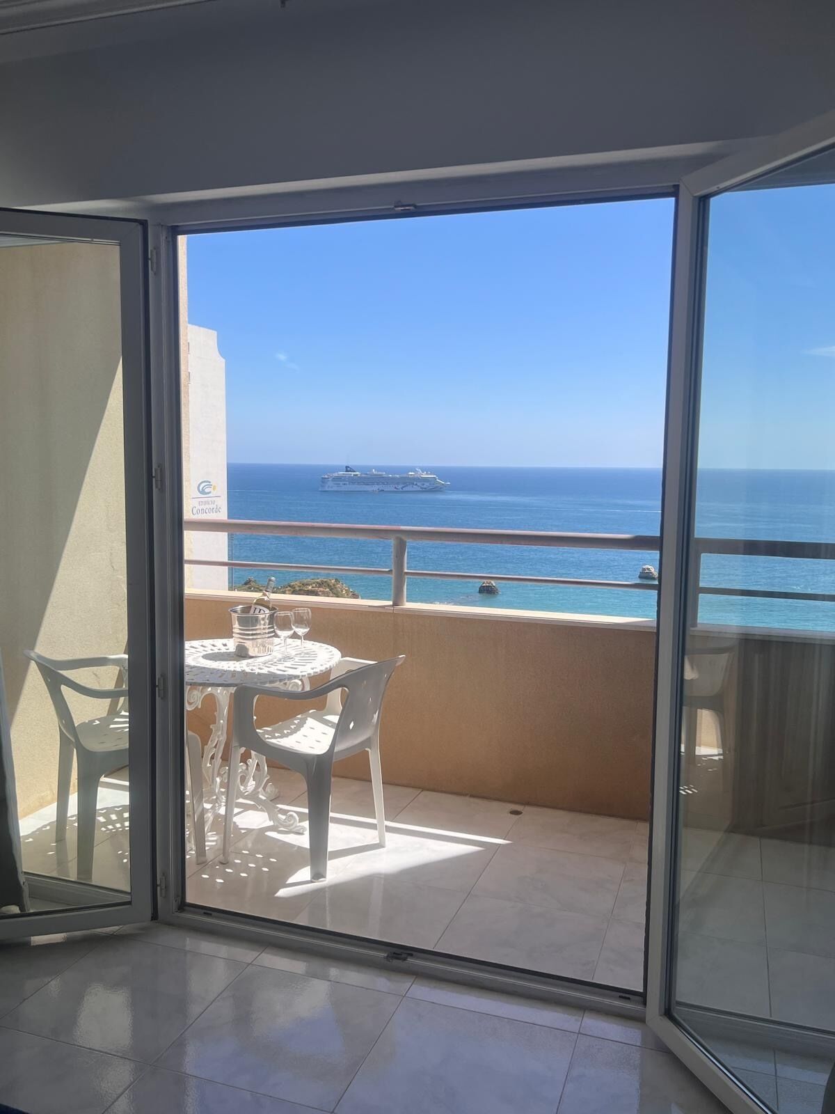 Uma varanda com mesa e cadeiras, vista para o mar e um navio ao longe.
