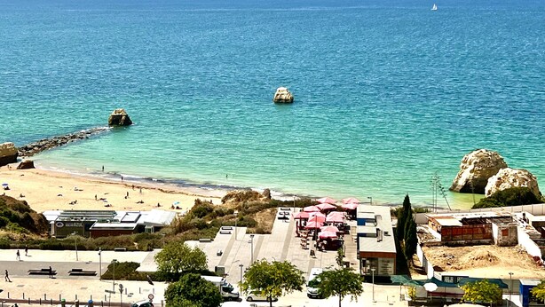 Une plage avec une eau turquoise et limpide, des affleurements rocheux et une rive sablonneuse. On y trouve des parasols et des tables installées pour les repas, et quelques personnes sont visibles sur la plage.