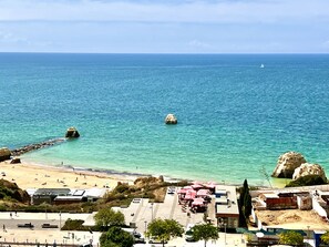 Une plage avec une eau turquoise et limpide, des affleurements rocheux et une rive sablonneuse. On y trouve des parasols et des tables installées pour les repas, et quelques personnes sont visibles sur la plage.