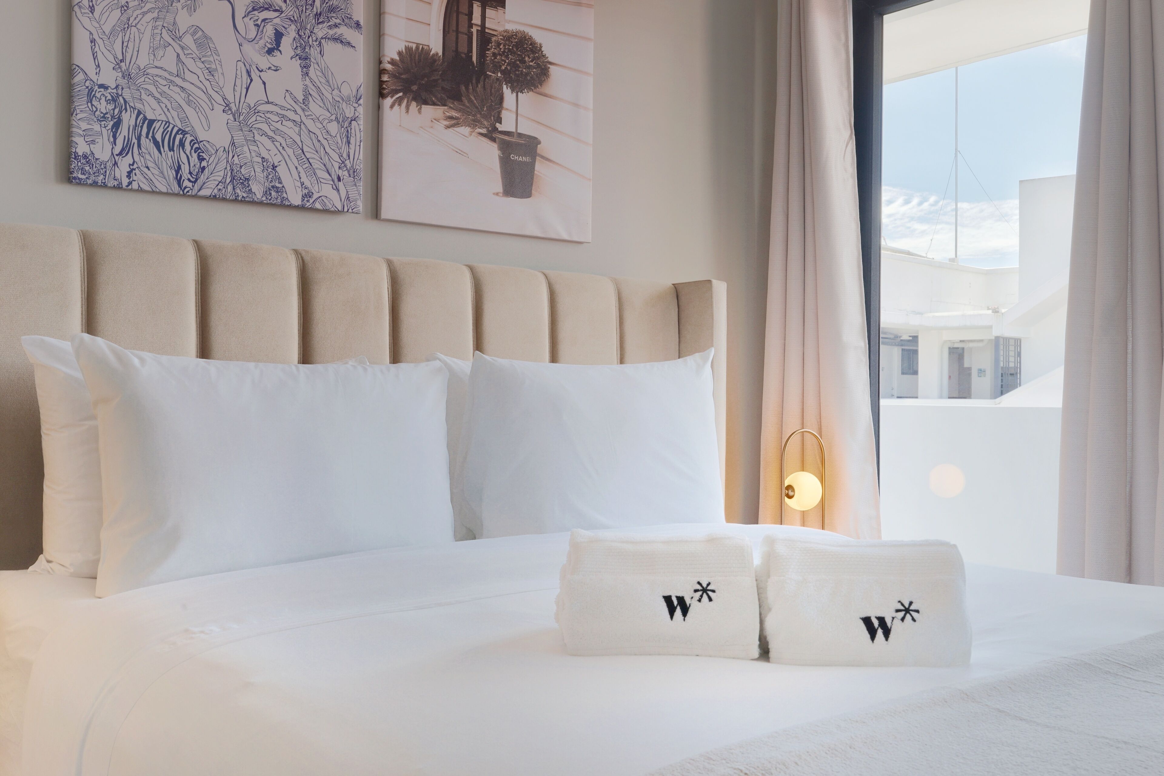 A hotel room with a large bed, white bedding, and two pillows with a logo. There is a window with curtains, a wall-mounted lamp, and artwork depicting a tiger and a plant.