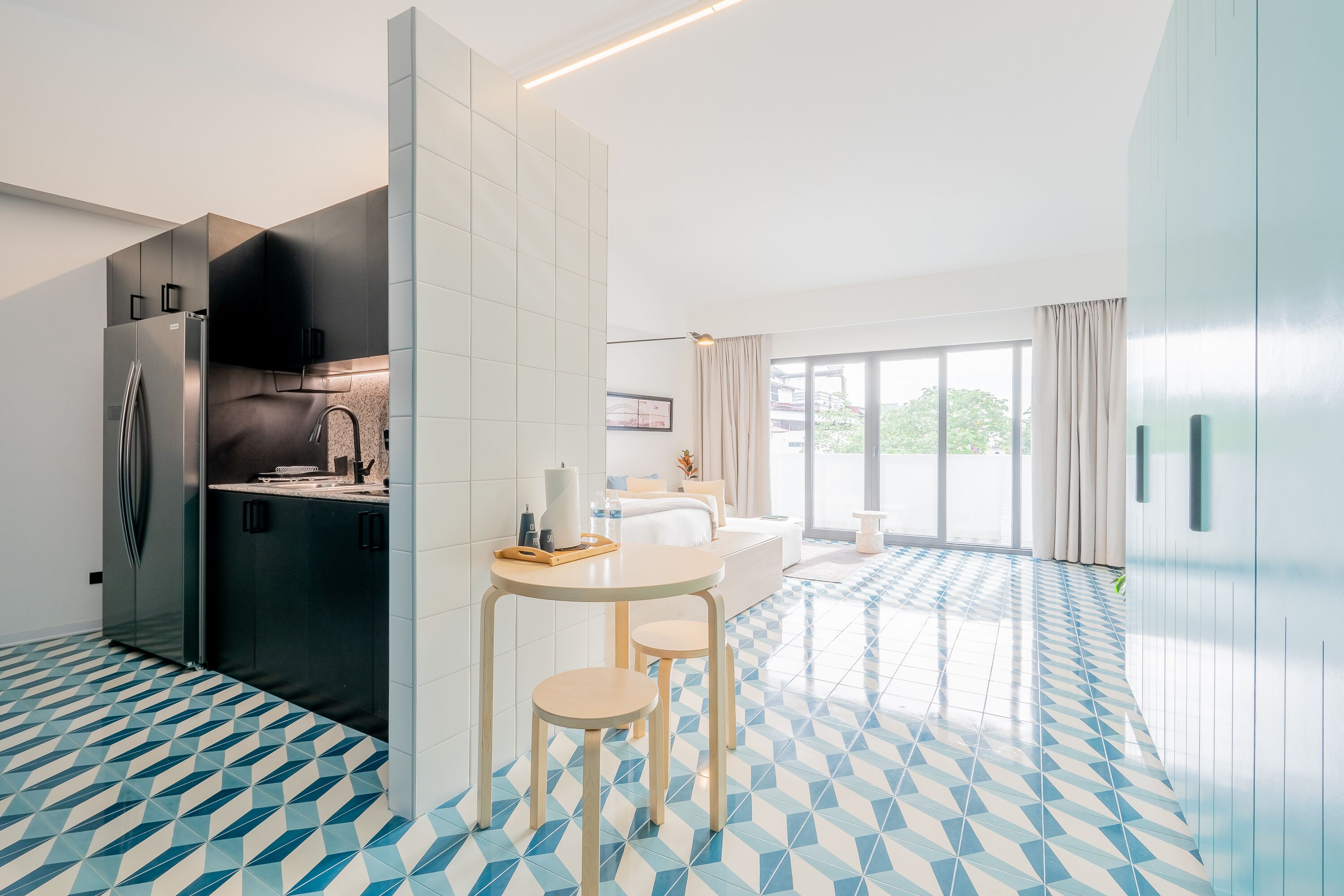 A modern kitchen with a built-in refrigerator, a dining area with a round table and two stools, and a large window providing natural light.