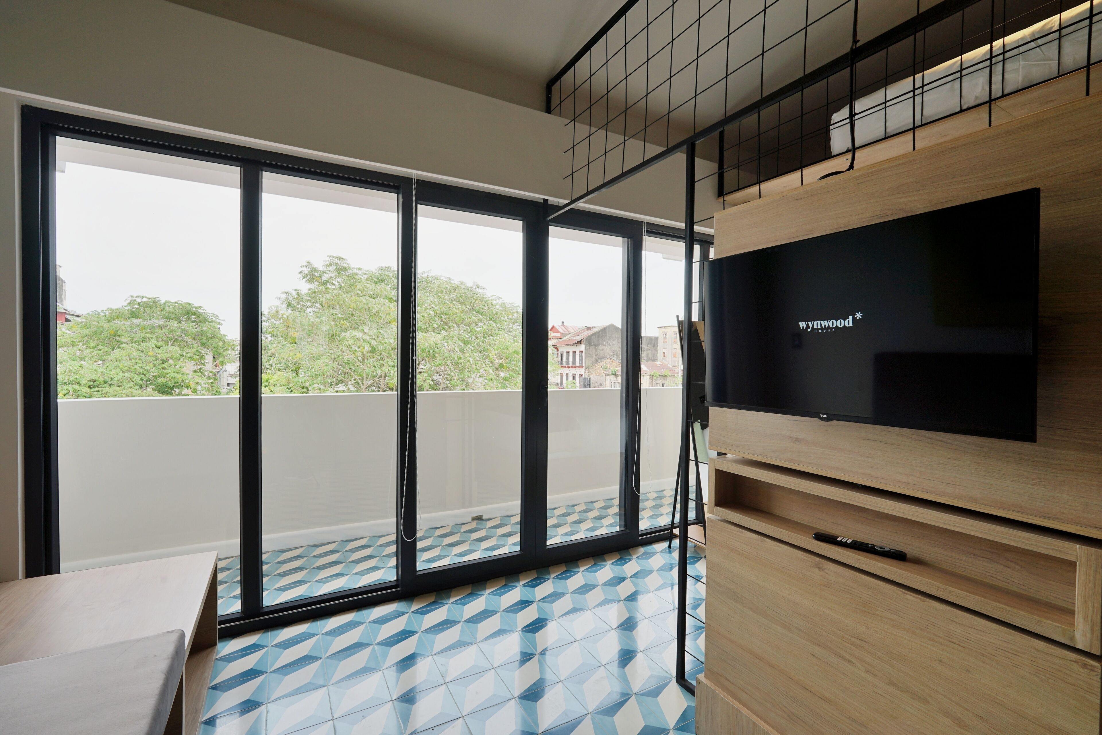 Ein modernes Zimmer mit einer großen Glasschiebetür, die auf einen Balkon führt, einem an einer Holzeinrichtung befestigten Flachbildfernseher und einem gemusterten Boden.