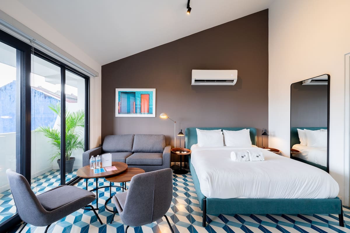 Ein modernes Hotelzimmer mit einem großen Bett, einer Couch, einem kleinen Tisch mit Wasserflaschen, einer Topfpflanze und einem großen Fenster mit Jalousien.