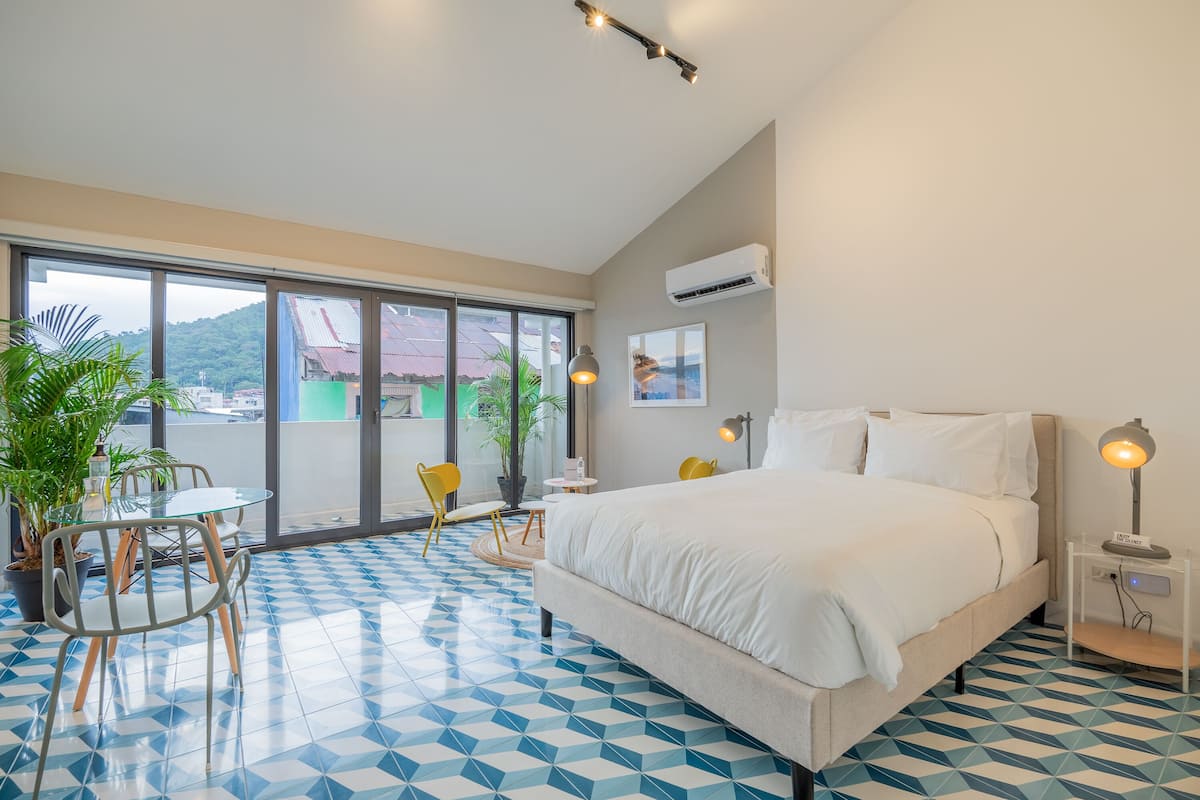 Ein Hotelzimmer mit einem großen Bett, einem Glastisch mit Stühlen, einer Topfpflanze und Blick ins Freie.
