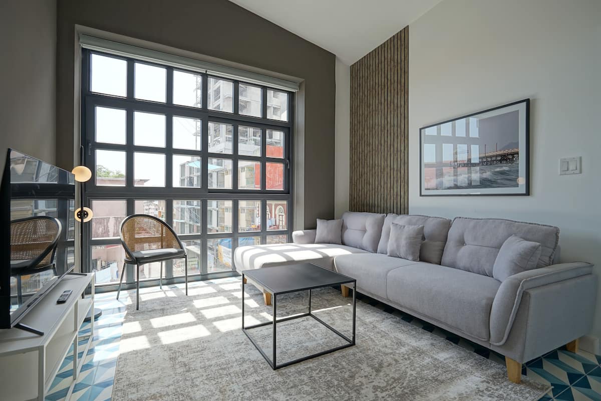Ein modernes Wohnzimmer mit einem großen Fenster, einem grauen Sofa, einem kleinen Tisch und einem Sessel.