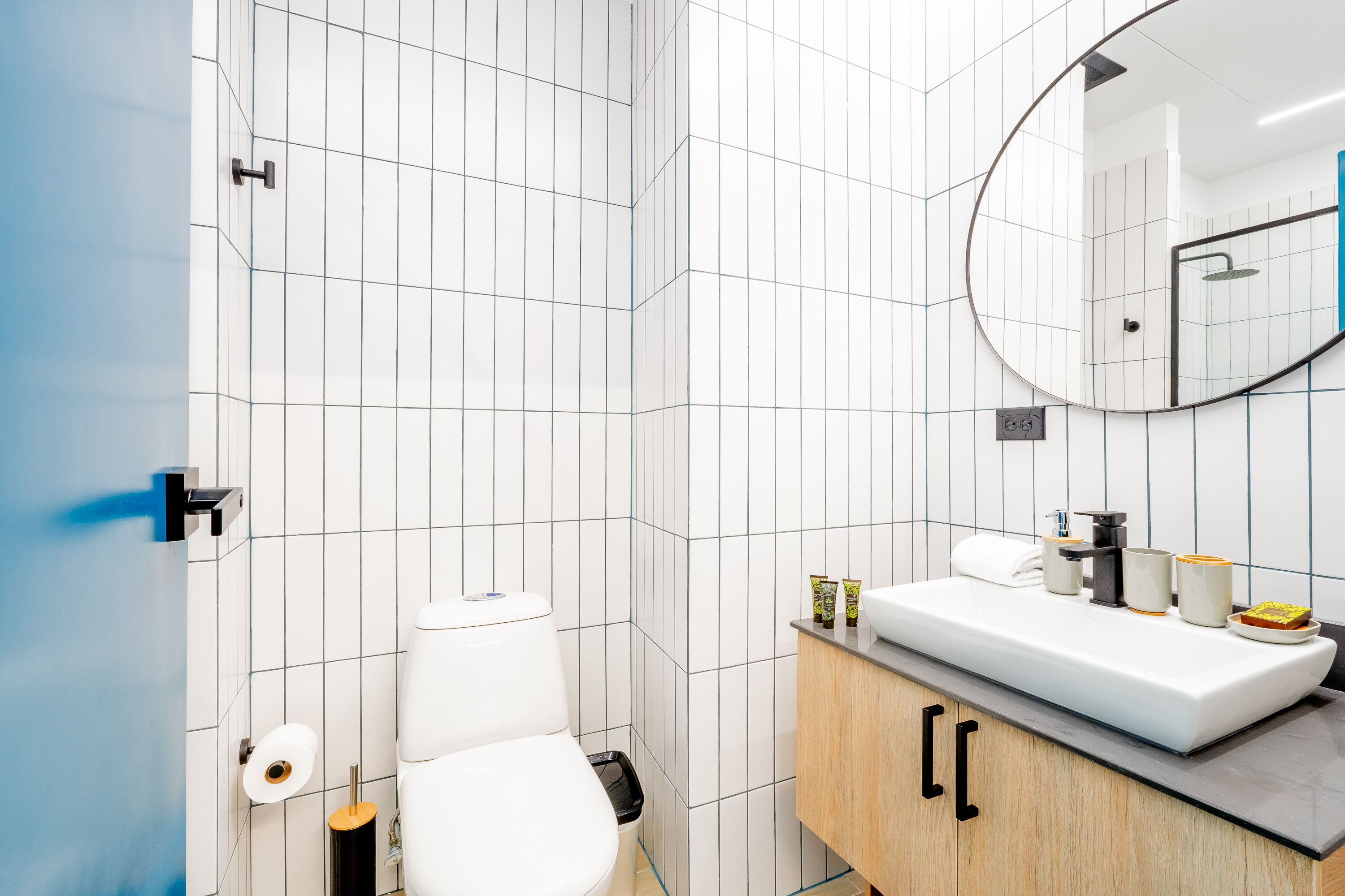 Un baño moderno con paredes de azulejos blancos, inodoro blanco, lavabo con espejo y un mueble de madera.