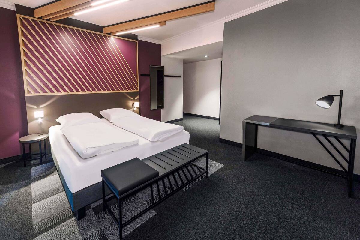 A modern hotel room with a bed, bedside tables, a desk, and a bench.