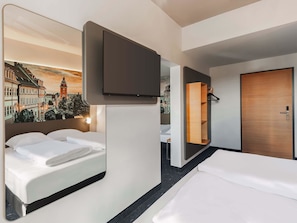 Habitación de hotel con dos camas, un televisor montado en la pared, una puerta de madera y un espejo que refleja la habitación.