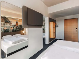 Habitación de hotel con dos camas, un televisor montado en la pared, una puerta de madera y un espejo que refleja la habitación.