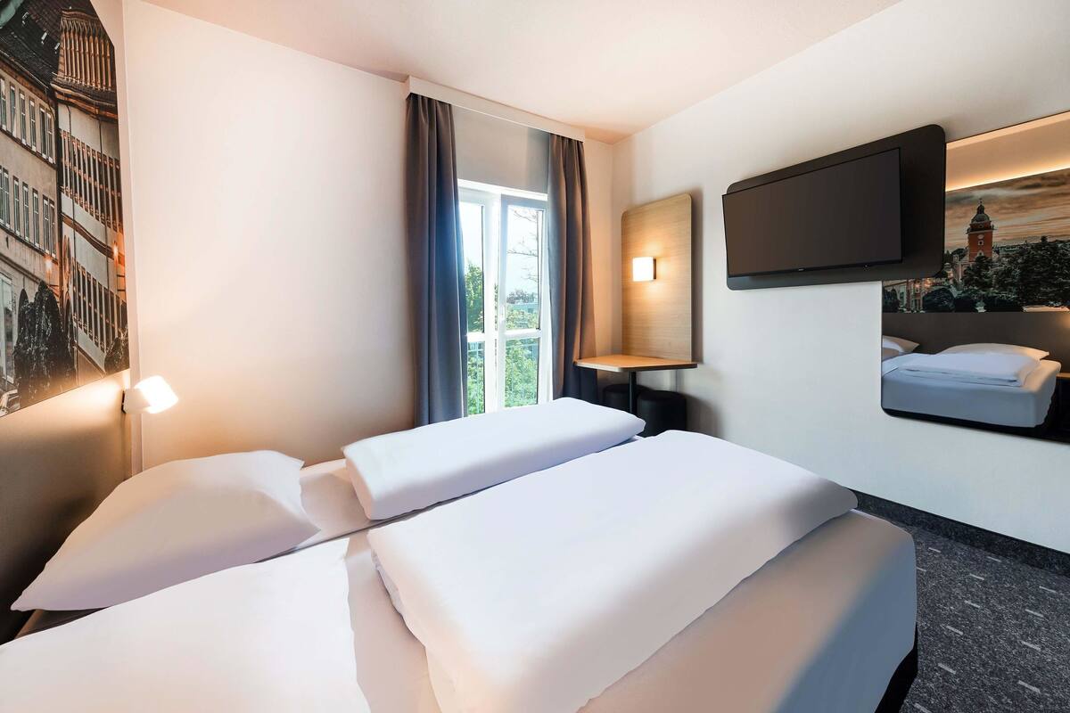 A hotel room with two beds, a TV mounted on the wall, a window with curtains, and a lamp.