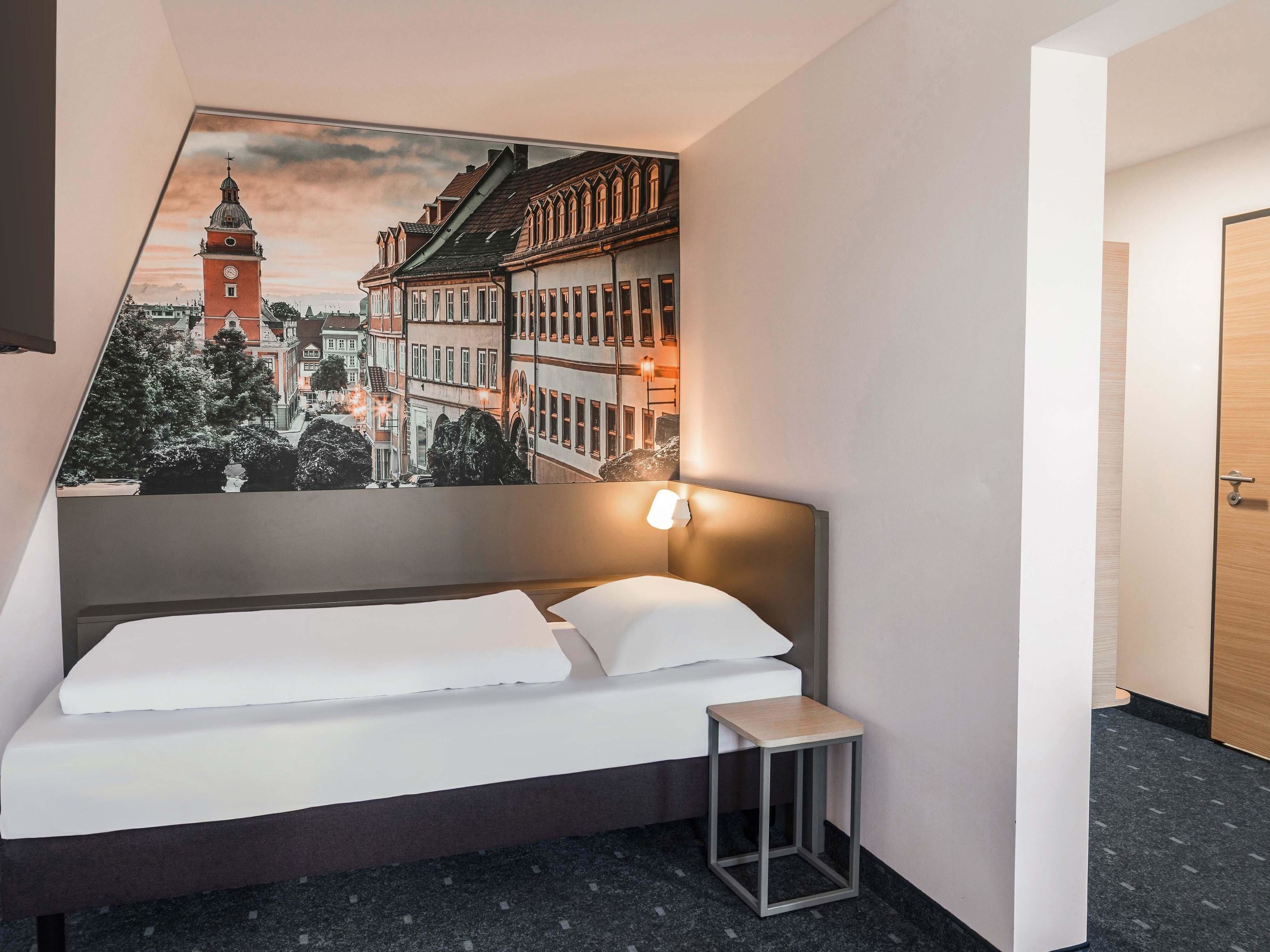 Ein Hotelzimmer mit Bett, Nachttisch, einem an der Wand hängenden Stadtbild und einer Tür.