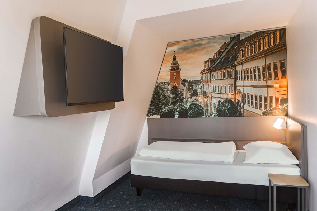 Habitación de hotel con una cama, un televisor montado en la pared, una mesita de noche pequeña y un cuadro de un paisaje urbano.