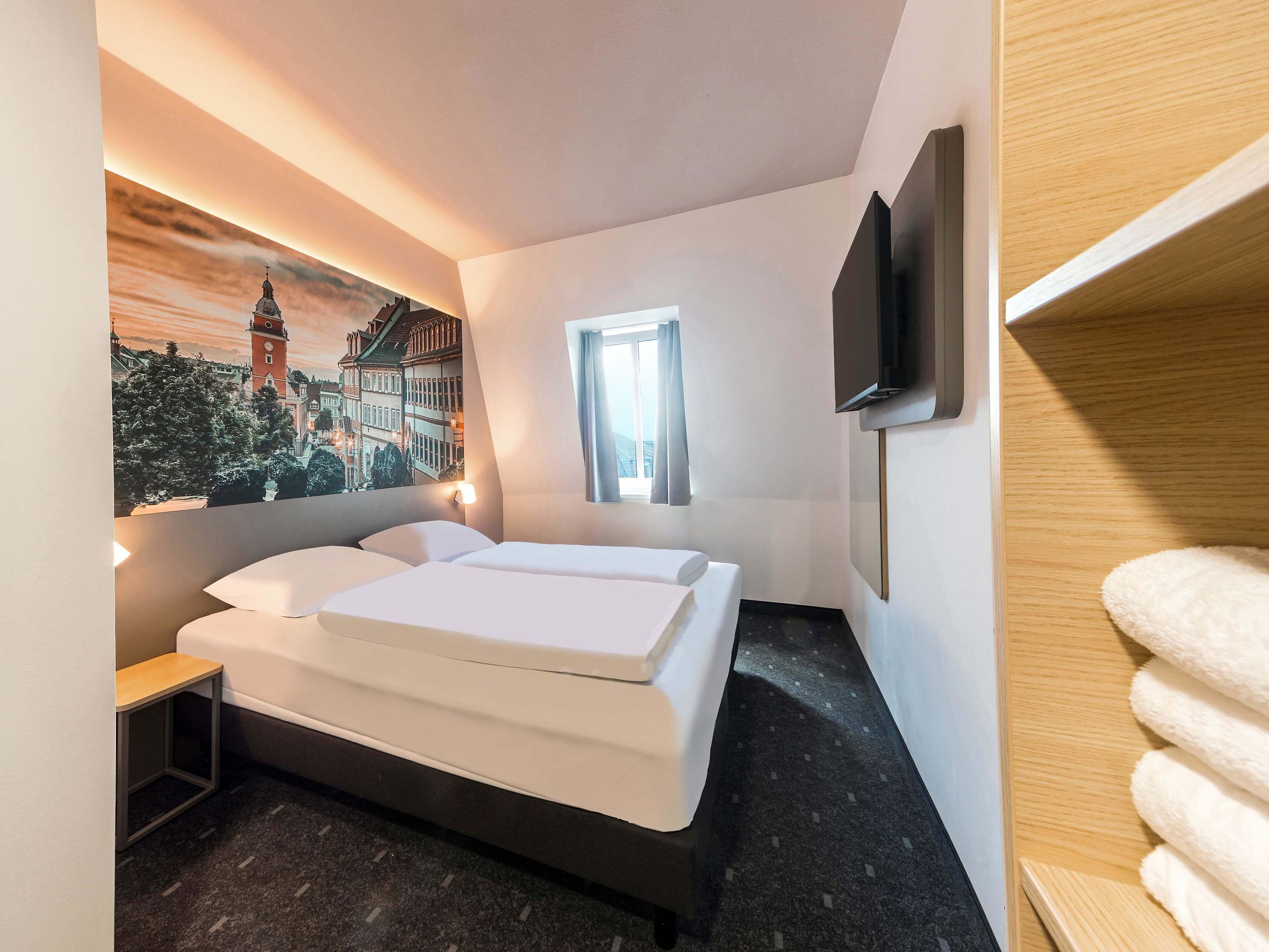 Una habitación de hotel con una cama, un televisor montado en la pared, una ventana con cortinas y un cuadro de un paisaje urbano.