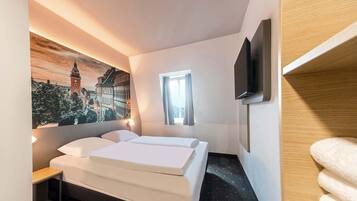 Una habitación de hotel con una cama, un televisor montado en la pared, una ventana con cortinas y un cuadro de un paisaje urbano.