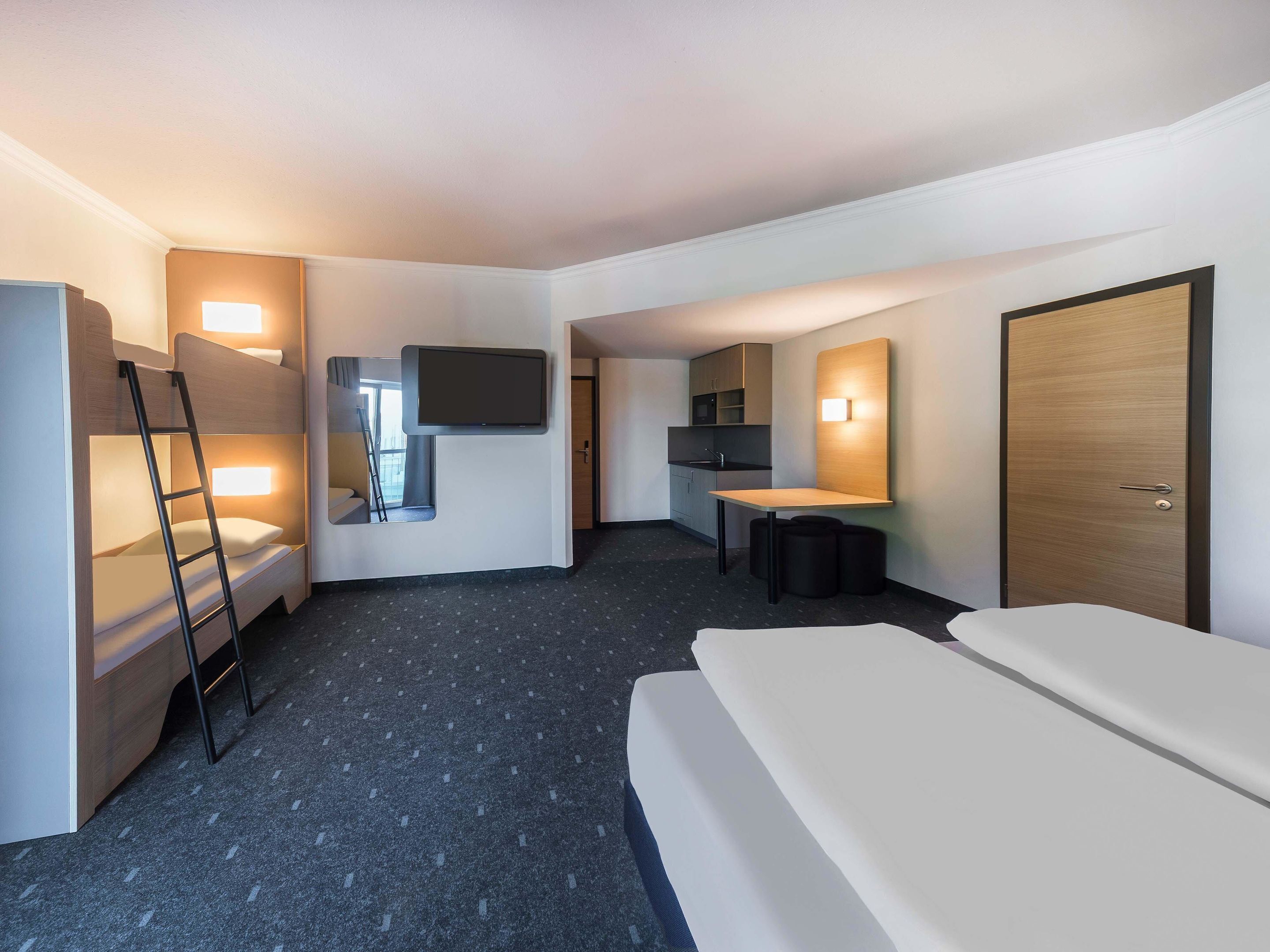 Ein Hotelzimmer mit Stockbetten, einem Schreibtisch, einem Flachbildfernseher, einer kleinen Küchenzeile und einer Tür.
