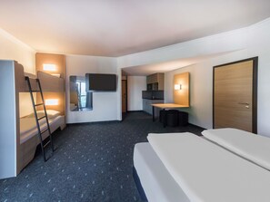Una habitación de hotel con literas, un escritorio, un televisor de pantalla plana, una kitchenette pequeña y una puerta.
