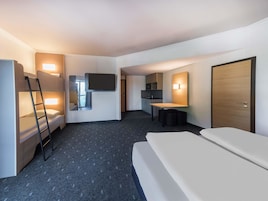Una habitación de hotel con literas, un escritorio, un televisor de pantalla plana, una kitchenette pequeña y una puerta.