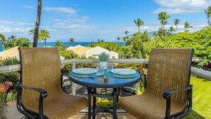 A balcony with two wicker chairs, a round table with a glass top, and a view of the ocean and palm trees. - Maui Banyan Kihei condo w/ large lanai and ocean views, summer deal (Kihei)