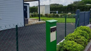 Ett inhägnat parkeringsområde med en grön teknikutrustningslåda, en byggnad med vit fasad och en häck längs trottoaren.