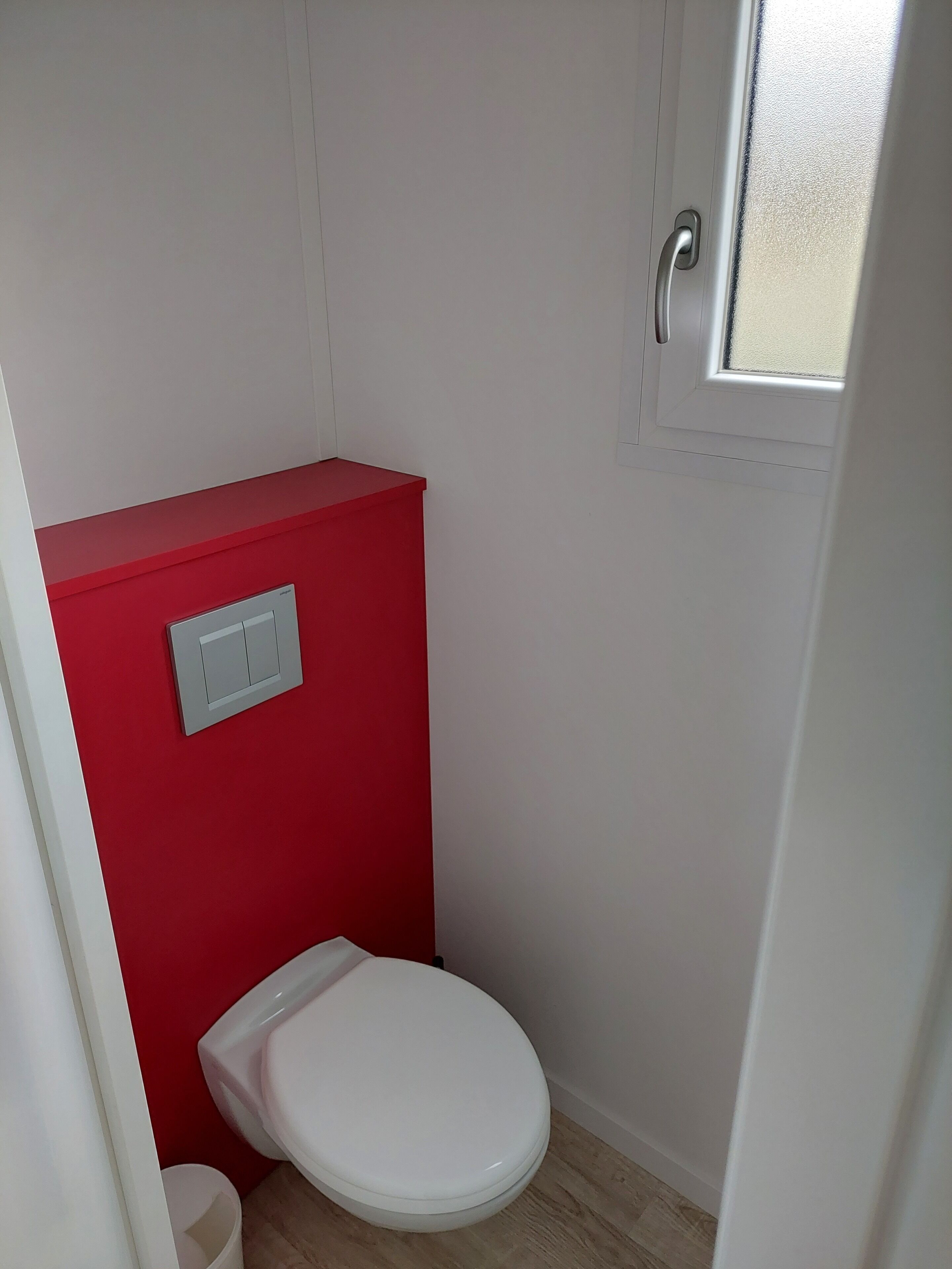A small, compact bathroom with a red accent wall, a white toilet, and a window.