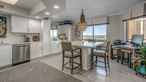 A modern kitchen with white cabinetry, a stainless steel dishwasher, and a dining area with a large island and bar stools. - Blue Ocean Front Escape (MYRTLE BEACH)