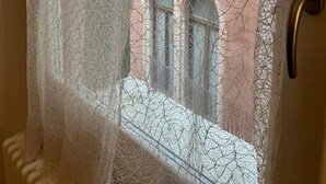 Une vue à travers un rideau de dentelle orné d’une poignée en métal, révélant un bâtiment aux fenêtres en arc de cercle et à la façade en briques.