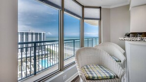 Un balcone con vista sull'oceano e una piscina, una sedia in vimini con cuscino a quadri, e un angolo cottura con un piano di lavoro e pensili.
