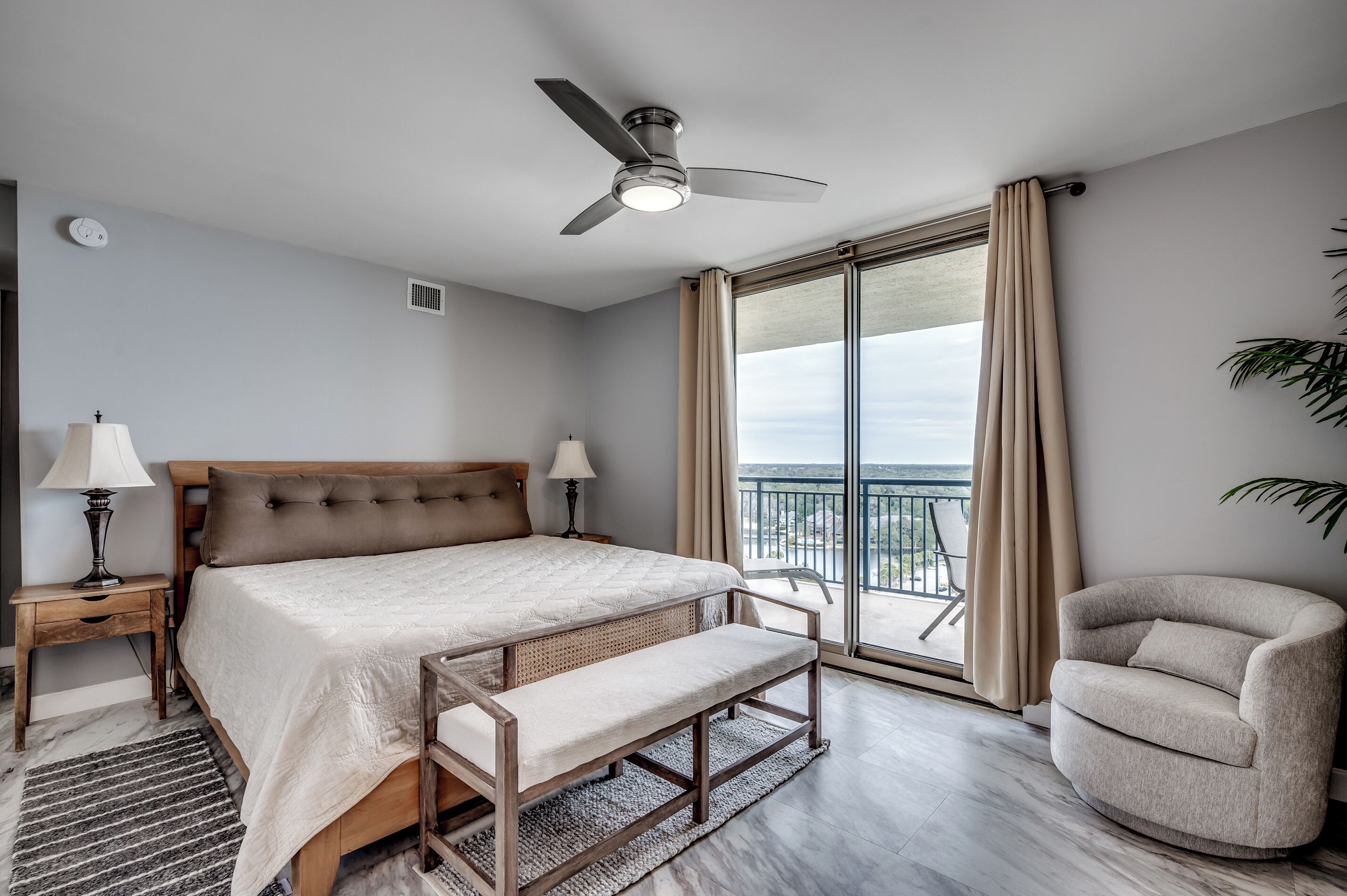 Un dormitorio con cama, mesitas de noche, un banco, ventilador de techo, balcón con una silla y vistas al exterior.