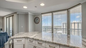 A modern kitchen with a marble countertop, white cabinetry, and a large window with blinds. - The Love Shack Myrtle Beach (Myrtle Beach)