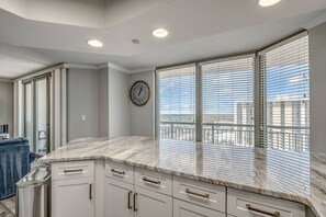 A modern kitchen with a marble countertop, white cabinetry, and a large window with blinds.