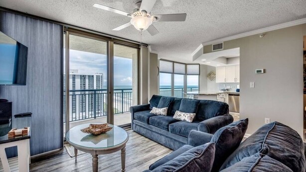 A spacious living room with a large sofa, a coffee table, and a ceiling fan. There is a large window offering a view of the city skyline. - The Love Shack Myrtle Beach (Myrtle Beach)