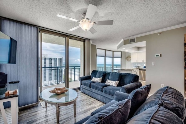 A spacious living room with a large sofa, a coffee table, and a ceiling fan. There is a large window offering a view of the city skyline. - The Love Shack Myrtle Beach (Myrtle Beach)