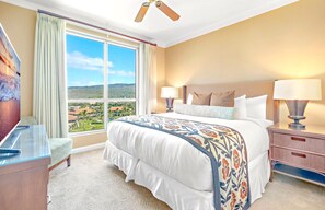 Ein Hotelzimmer mit einem großen Bett, zwei Nachttischlampen, einem Nachttisch, einem Schreibtisch und Blick auf eine Landschaft durch große Fenster.