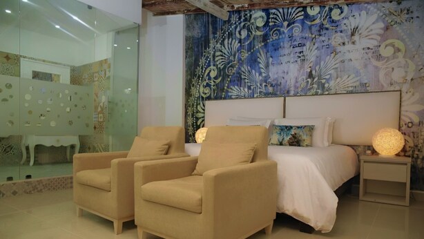 A bedroom with a bed, two armchairs, a nightstand with a lamp, and a decorative tapestry on the wall. - HOTEL HABANA REAL (Apartadó)