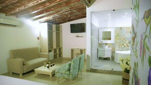 Habitación de hotel con sofá, una silla, televisor, baño con espejo y suelo de baldosas.